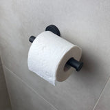 Un rouleau de papier toilette est suspendu au porte-rouleau de papier toilette Tino Black de yessential.fr, monté contre un mur de salle de bain carrelé gris clair pour une touche moderne.