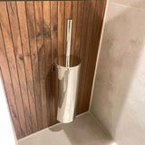 Le porte-brosse de toilette Branco Chrome de yessential.fr a une poignée argentée brillante, fixée au mur contre des panneaux de bois verticaux à côté des murs et du sol carrelés beige clair.