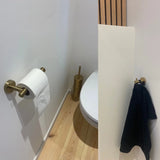 Une petite salle de bain moderne avec un parquet en bois clair, des toilettes blanches, un porte-papier toilette et une brosse dorés, et le crochet Justo Gold de yessential.fr retenant une serviette noire pour une touche élégante.