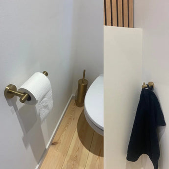 Une petite salle de bain moderne avec un parquet en bois clair, des toilettes blanches, un porte-papier toilette et une brosse dorés, et le crochet Justo Gold de yessential.fr retenant une serviette noire pour une touche élégante.