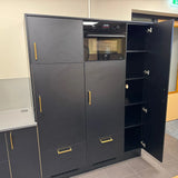 Les armoires de cuisine hautes noires Vasco Gold de yessential.fr ont des poignées dorées et un four intégré. Une porte est ouverte, exposant plusieurs étagères. Les armoires frappantes contrastent magnifiquement avec le mur et le sol de couleur claire.