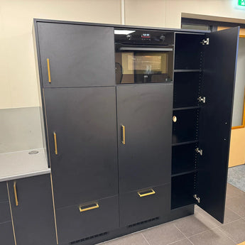 Les armoires de cuisine hautes noires Vasco Gold de yessential.fr ont des poignées dorées et un four intégré. Une porte est ouverte, exposant plusieurs étagères. Les armoires frappantes contrastent magnifiquement avec le mur et le sol de couleur claire.