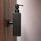 Le distributeur de savon Sonego Black de yessential.fr est monté sur un mur en bois, avec une finition noire mate moderne, un design minimaliste et une petite fenêtre pour vérifier facilement le niveau de savon.