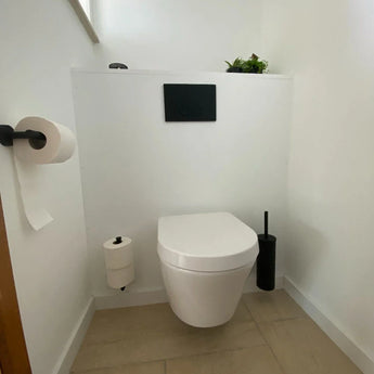 Une salle de bain moderne et minimaliste avec des toilettes blanches suspendues, l'ensemble WC Lorenzo Black yessential.fr (comprenant une brosse et un support de toilettes noirs), deux rouleaux de papier toilette sur supports et une petite plante sur une étagère contre des murs blancs et un sol carrelé clair.