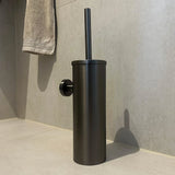 L'ensemble de toilette Mano Gunmetal de yessential.fr - une brosse de toilette noire avec un support cylindrique - répond aux carreaux clairs de salle de bain près du mur, qu'une serviette beige est suspendue à l'arrière-plan, avec une touche sophistiquée.