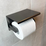 Un rouleau de papier toilette blanc sur le support yessential.fr Tazio Gunmetal (No Drill) avec un dessus plat, monté sur un mur de salle de bains carrelé gris clair.