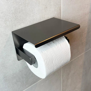 Un rouleau de papier toilette blanc sur le support yessential.fr Tazio Gunmetal (No Drill) avec un dessus plat, monté sur un mur de salle de bains carrelé gris clair.