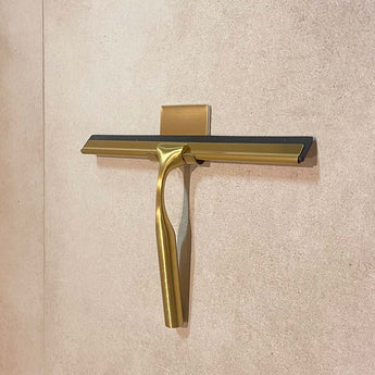 Le Zamoro Gold de yessential.fr, doté d'une poignée de douche de couleur dorée avec une lame en caoutchouc noir, est monté sur un mur carrelé beige dans un support doré assorti.