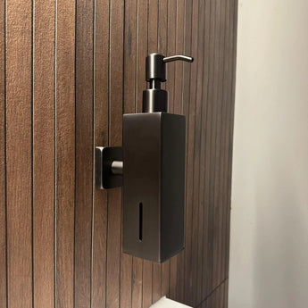 La pompe à savon Sonego Gunmetal de yessential.fr, en noir élégant avec des accents en acier inoxydable, est fixée au mur sur un mur à lattes en bois dans la salle de bain pour un look moderne et élégant.